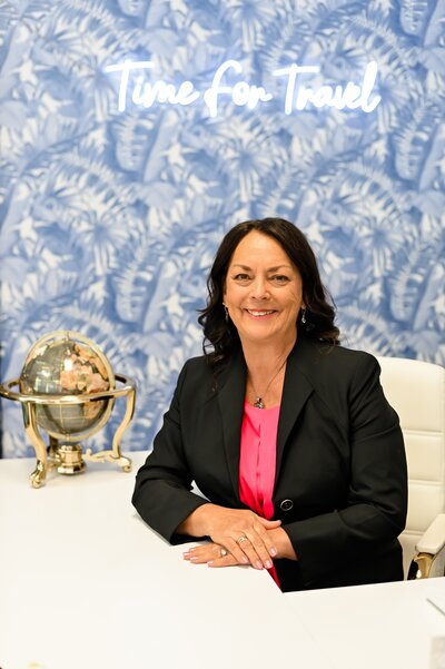 A smiling travel agent in a black blazer and pink blouse, specializing in affordable destination wedding planning and honeymoon trips.
