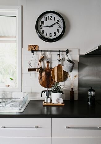 organized kitchen countertop with a black and white clock hanging over a rod holding hanging cutting boards over a coffepot and plant sitting on a black countertop with a white dish drying rack to the left in front of an open window