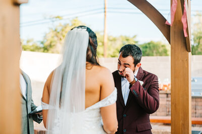 A photo of a groom wiping a tear while crying during his wedding ceremony as the bride looks on
