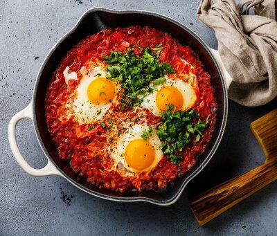 A close-up of fertility-boosting traditional shakshuka served in a white-handled skillet, featuring vibrant poached eggs with bright yellow yolks nestled in a rich, spiced tomato sauce. Freshly chopped parsley is sprinkled on top, adding a pop of green for colour and nutrients. The pan rests on a textured grey surface with a rustic beige linen cloth nearby, creating a warm, homely scene.
