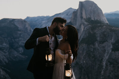 groom kissing bride on the cheek while she smiles they are both holding up lanterns behind them is half dome