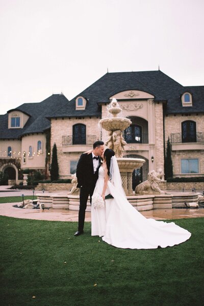 The scene is composed with the couple centered, positioned slightly to the left of the image, in front of a large, elaborate house and a fountain