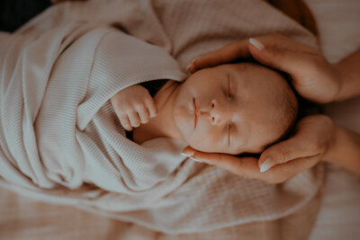 A mother's hands cradle her sleeping baby's head as he peacefully lays on his back swaddled in a cream colored wrap.