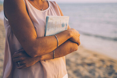 A woman stands on the beach, holding a book and enjoying the sun and waves in the background.