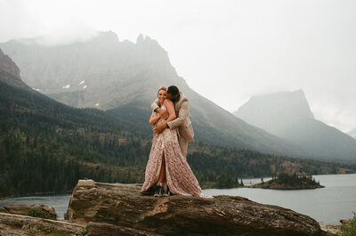 Couple embraces on a rocky cliff during their elopement in Glacier National Park with misty mountains, forested slopes, and a serene lake in the background.
