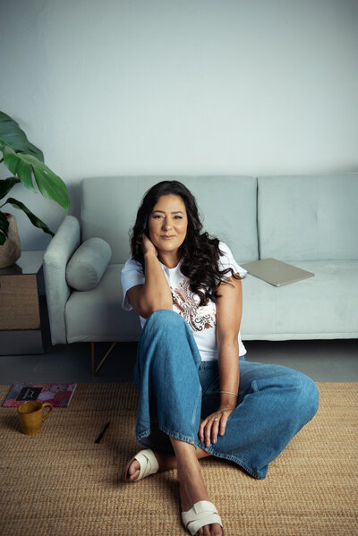 Asmahan, online business manager, sitting thoughtfully on the floor of her home office in casual jeans and white tee, reflecting her approachable leadership style