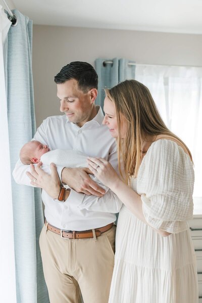 A couple stands by the window cradling their newborn baby during their in -home NJ newborn photo session