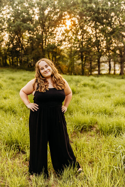 beautiful senior girl posing in a field and looking super confident in her black jumpsuit by Ashley Kalbus