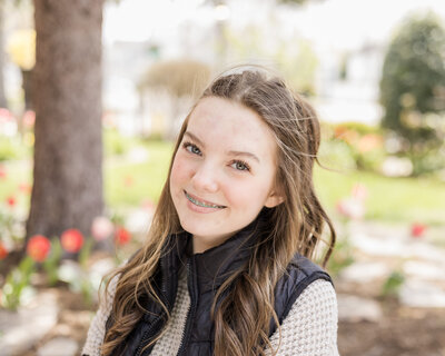 high school senior female, with long brown hair, wearing a black zip up jacket over a light tan long sleeve shirt, looking at the camera, smiling