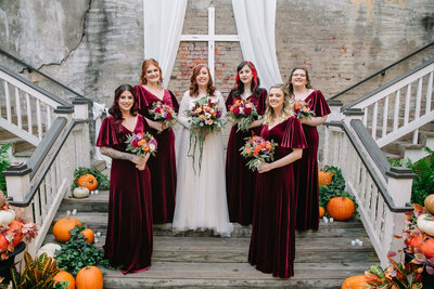 A bride in a white dress stands with five bridesmaids in matching burgundy dresses, all holding bouquets, on wooden stairs decorated with pumpkins and greenery. A white cross and drapes are displayed behind them.
