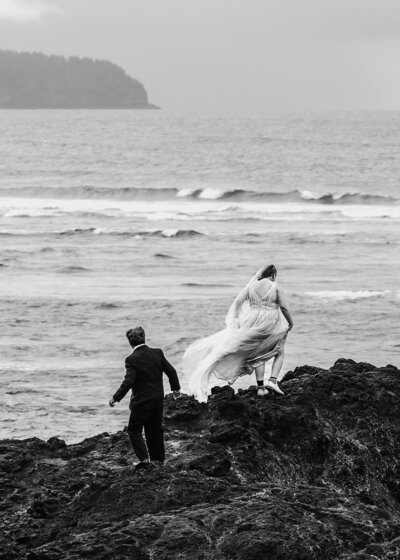 during their full day elopement package, a couple explores a rocky beach. The Bride leads, her dress billowing in the wind as her groom follows. 