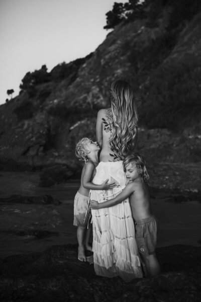 A woman with long hair and a tattoo on her back stands on a rocky shore, facing away. Two young shirtless children stand by her sides, holding onto her. They are all barefoot and appear relaxed.