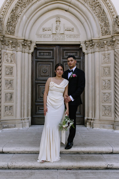 Bride and groom standing in front of a building with straight faces.