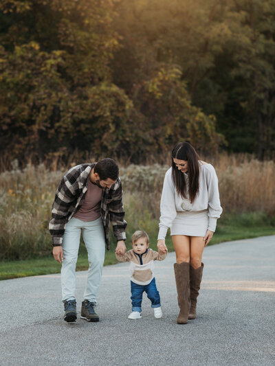 mom and dad holding one year old sons hand walking in park for family photos