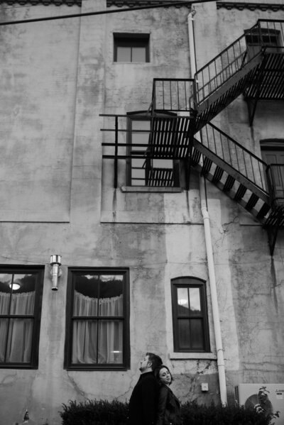 couple standing back to back in front of tall building with large windows and fire escape stairs