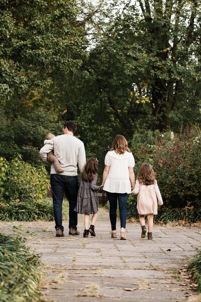A family of five, captured by a Pittsburgh family photographer, walking away from the camera on a garden path, with the father carrying a small child on his shoulders.