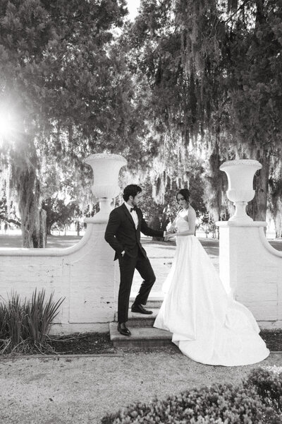 Black-and-white photo of a bride in a flowing gown and a groom in a suit, standing together by a stone balustrade under tall trees with Spanish moss.