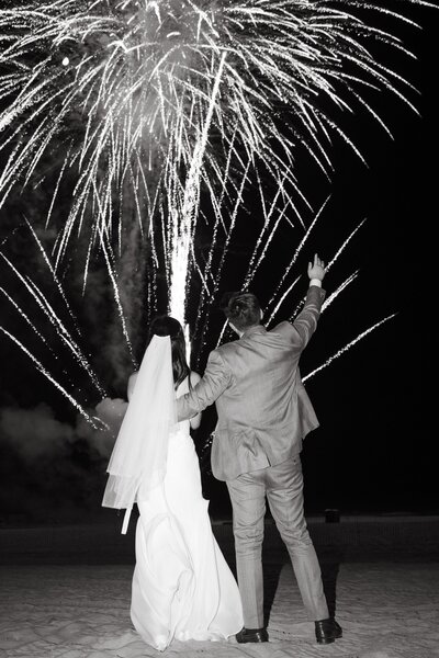 Two newlyweds in formal attire stand on a beach at night, watching a large display of fireworks in the sky during their wedding reception at Hotel del Coronado. This wedding was planned and designed by Thater Made, a luxury wedding and event producer based in San Diego, CA.