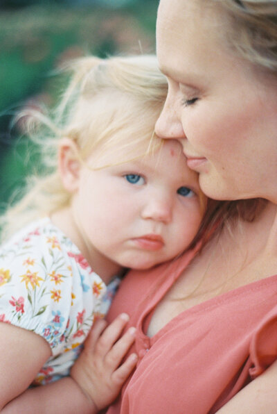 Toddler girl in floral dress snuggles into mom's chest with somber face while mom gently smiles with eyes closed wearing coral dress by Portland Family Photographer Emilie Phillipson Photography. 