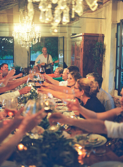Wedding guests raising glasses for a toast during a warm, intimate reception dinner under chandeliers with a live musician playing in the background.