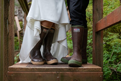 Close-up of a bride and groom wearing Xtra Tuff boots on a wooden bridge, with the bride’s white wedding dress flowing above the boots. The setting is lush and green, capturing the adventurous spirit of an Alaskan elopement in the wilderness.