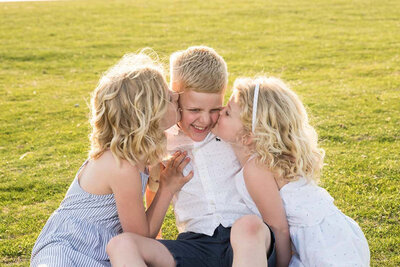 Three smiling children with blond hair sit on grass. Two girls in dresses kiss a boy in a white shirt on his cheeks. The scene is joyful and sunny.