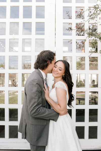 Groom wearing gray suit hugging bride wearing wedding dress with diamond detailing on shoulder in front of white barn doors.