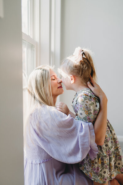 mom touching noses with little girl by a window