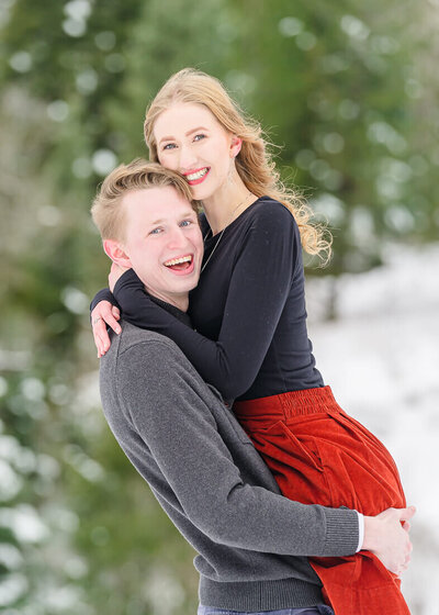 Utah engagement photography of a man wearing a dark blue polo shirt with his arm around a woman  in a white lace dress celebrating their engagement in front of a blossoming tree in American Fork