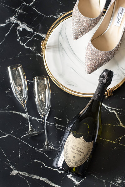 A pair of glittery high heels is placed on a round mirror. Beside them, there are two empty champagne flutes and a bottle of Dom Pérignon champagne on a black marble surface.
