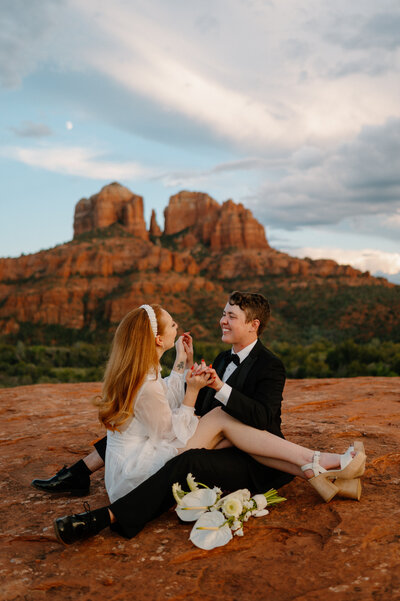 A wedding ceremony in Sedona, Arizona, where the couple exchanges vows under a beautifully decorated triangular wooden arch. The officiant stands between the marriers, who hold hands and smile at each other, with red rock formations in the background.