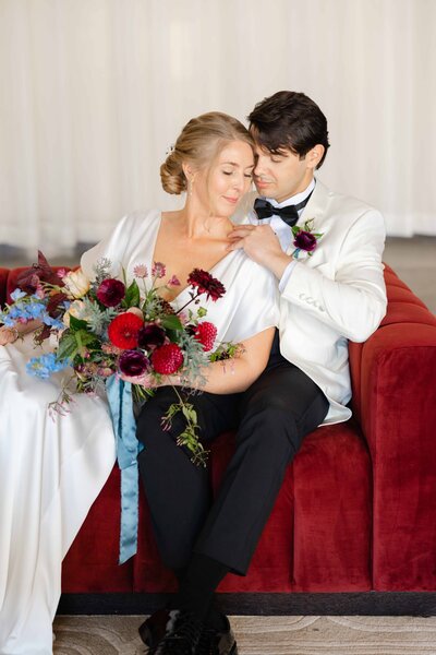Bride and groom sit closely on a deep red velvet couch, sharing an intimate moment. The bride, in a sleek white gown, holds a vibrant bouquet of red, purple, and blue florals with trailing ribbons. The groom, dressed in a white tuxedo jacket with a dark bow tie, gently touches her shoulder as they lean into each other, radiating love and elegance.