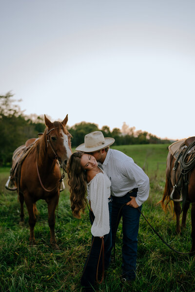 man kissing woman while standing in a field next to their horses