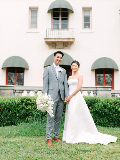 A smiling couple is in wedding attire in front of muckenthaler mansion building with green awnings. The bride holds a bouquet of white flowers. The scene is joyful and elegant.