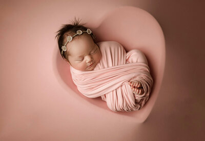 Newborn baby girl swaddled in blush pink, peacefully sleeping in a heart-shaped bowl on a matching soft pink background, symbolizing warmth and love captured in Minneapolis studio by Duehr Photography