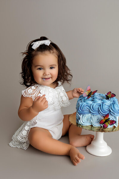 A smiling baby sits on the floor next to a blue frosted cake adorned with colorful butterfly decorations. The baby is wearing a white lace outfit with a matching headband, looking adorable and happy. The background is a soft, neutral gray, which keeps the focus on the baby's joyful expression and the vibrant cake.