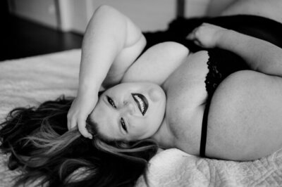 empowering boudoir photography with woman in black lingerie lays on a white sheet bed as she smiles into the camera