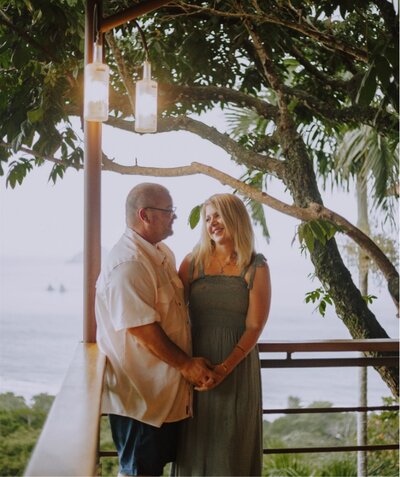 Couple holding hands and smiling on a balcony surrounded by lush tropical trees and ocean views at sunset.