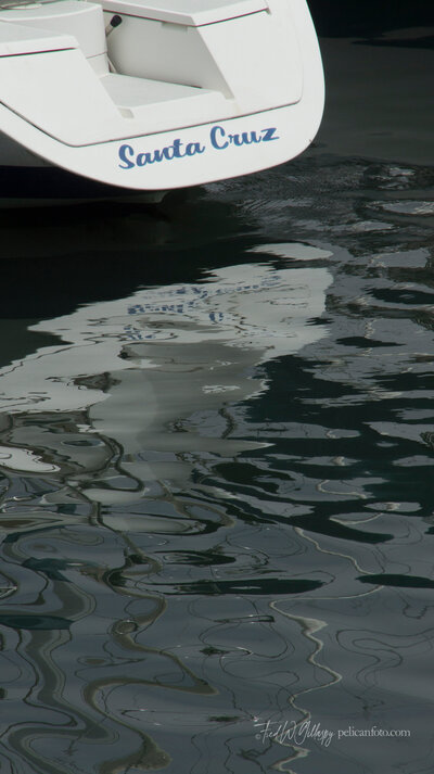 Stern of a sailboat adorned with the words “Santa Cruz,” reflected in the waters of Santa Cruz Harbor, CA.
