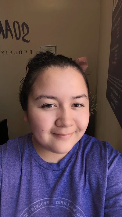 A person with dark hair in a bun smiles at the camera while wearing a purple shirt. The soft, natural lighting highlights a cozy space in Tolleson, Arizona—ideal for massage therapy—with beige walls and partially visible text and a framed item.