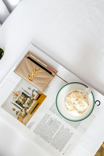 Cup of cappuccino, lifestyle magazine, and YSL cardholder styled on white bedding—evoking luxury and intentionality