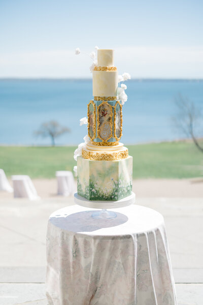 bespoke wedding cake with fine art painting and gilded age details hand painted on in front of a backdrop of Narragansett bay