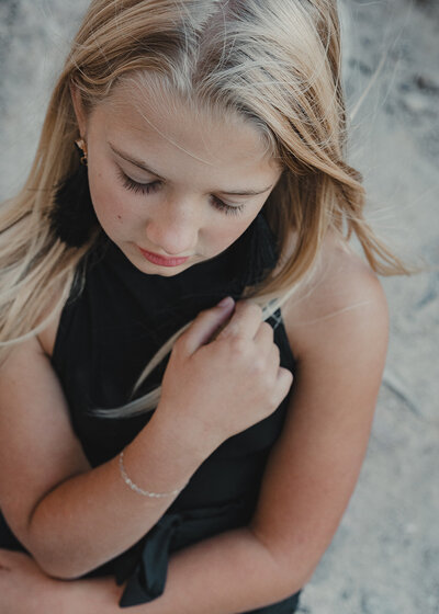 A preteen, blonde girl how's the wind blowing in her hair as her right arm caresses her left shoulder and she looks down words while having a thoughtful expression.