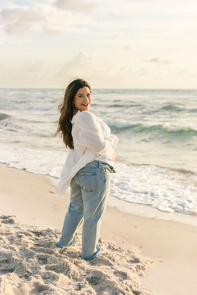 Smiling pregnant woman in white shirt and jeans during sunrise maternity session on Miami beach.