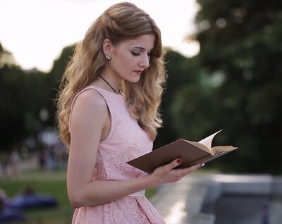 Young female romance novelist reading book in park, showcasing inspiration for fiction writers and aspiring authors in publishing industry