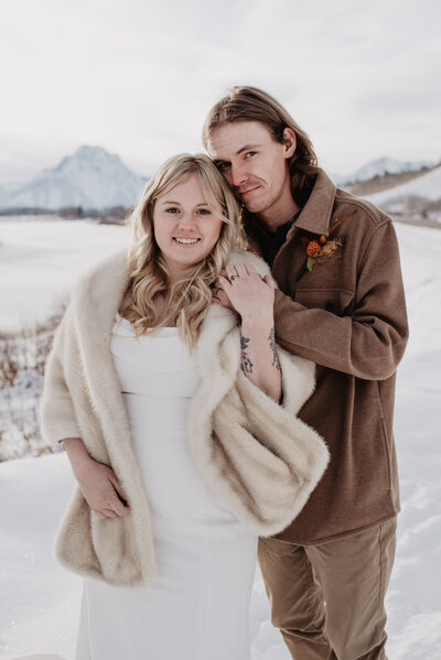 Grand Teton wedding with bride in a faux fur coat over her wedding dress with her groom standing next to her shoulder and leaning into her captured by Jackson Hole photographers