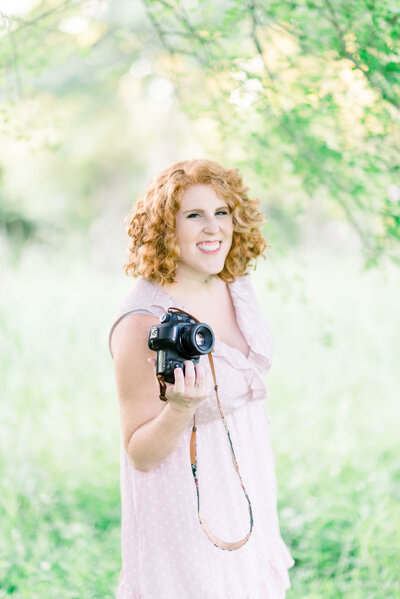 Woman with shoulder-length curly red hair wearing pink dress with ruffles and small white polka dots and holding a black camera with brown camera strap and standing in area of greenery taken by New Orleans wedding photographer Elizabeth Collins