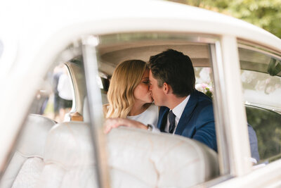 Inside a vintage car parked at New Barn Farm, a couple shares a tender kiss. The woman with blonde hair leans in towards the man wearing a blue suit. Sunlight streams through the windows, casting a warm glow on the beige interior, while Seer Green's serene countryside surrounds them.