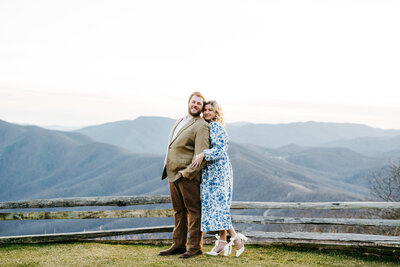 engagement photos by Virginia wedding photographer with man in a brown suit standing on an outlook in Shenandoah national park with a woman in a white and blue dress standing behind him and holding his arms with the mountains behind them as they tilt their heads towards each other