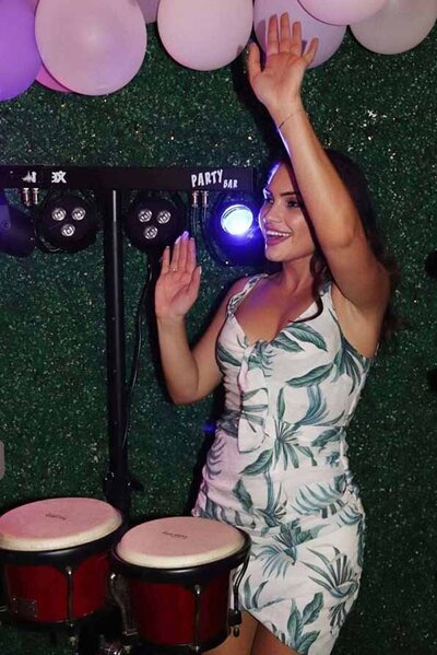 Female percussionist in a tropical dress raising her hands while playing bongos at a vibrant party with pink balloons and lights.